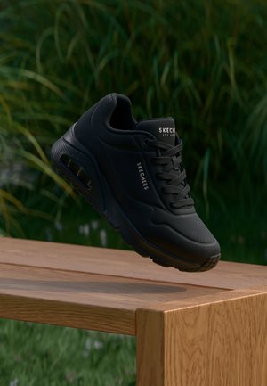 Sort Skechers sneaker med synlig luftpolstret sål, svævende over en træbænk mod en grøn plante baggrund udendørs.