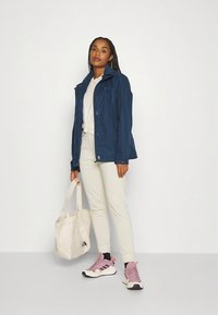 Tamsiai mėlynas vėjas, šviesiai pilka marškinėliai, smėlio spalvos treningai, rožiniai ir balti sportbačiai, laikantis smėlio spalvos pirkinių maišelį. Švelni medžiaga, laisvas siluetas.