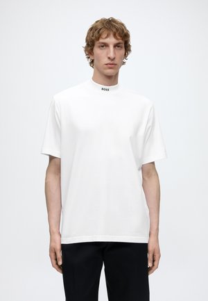 Jeune homme aux cheveux bouclés portant une chemise blanche à manches courtes avec le logo « BOSS » sur un col rond haut, associée à un pantalon noir.