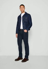 Chaqueta de mezclilla oscura con cierre de botones y dos bolsillos en el pecho, llevada sobre una camiseta blanca y jeans oscuros, combinada con zapatos marrones.