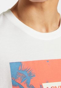 T-shirt en coton blanc avec un col rond, ornée d'une impression graphique corail et bleue, comprenant des feuilles de palmier et du texte au centre.