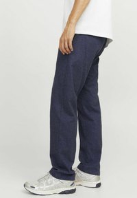 Pantaloni blu scuro con tessuto testurizzato, taglio dritto e tasche, abbinati a scarpe da ginnastica argentate e una camicia bianca.