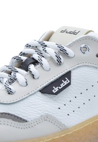 Sneaker bianco e beige con lacci bianchi a strisce nere, etichetta con logo nera, dettagli perforati e suola in gomma visibili da un primo piano.