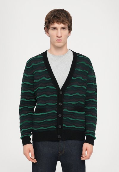 Zwarte cardigan met groene golvende strepen, vijf knopen en geribbelde manchetten. Gelaagd over een grijs shirt en heeft twee voorkruiden.