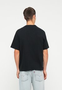 Sort bomulds t-shirt med afslappet pasform, korte ærmer og rund halsudskæring. Båret med lyseblå denim jeans, set fra bagsiden.