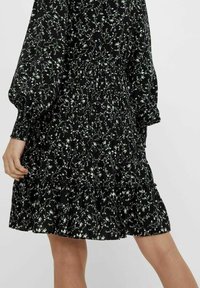 Robe noire avec imprimé floral blanc, présentant des manches bouffantes et un ourlet à volants superposés. Fabriquée en tissu léger, avec un détail de taille élastique.