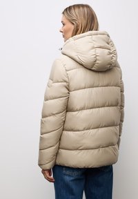 Giacca puffer beige con cappuccio, caratterizzata da sezioni trapuntate e polsini elastici. Tessuto liscio con una leggera lucentezza; dettaglio del logo discreto sulla manica.