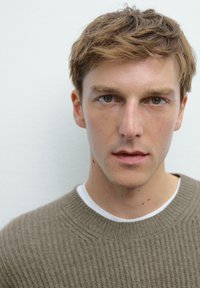 Bruine gebreide trui gedragen over een wit shirt; close-up van een persoon met kort, rommelig haar en bruine ogen tegen een neutrale achtergrond.