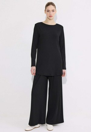 Kobieta w czarnej tunice z długim rękawem i szerokich czarnych spodniach stojąca na jednolitym białym tle w białych trampkach.