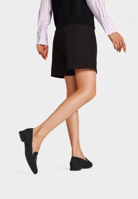 Mocassini neri con punta rotonda, una tracolla decorativa e una fibbia, e un tacco basso, abbinati a pantaloni shorts neri e una camicia a righe.