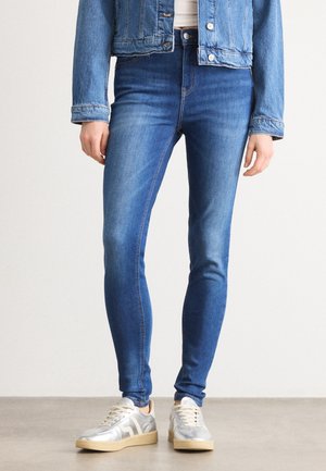Person trägt blaue Skinny-Jeans, eine passende blaue Denimjacke, weißes Hemd und silberne Sneakers mit beigen Sohlen und steht auf einem grauen Boden.