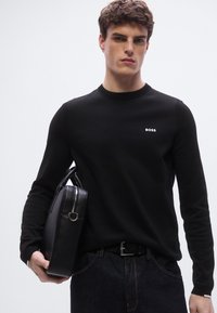 Musta pitkähihainen paita, jossa on resorinen pääntie ja valkoinen "BOSS"-logo, yhdistetty tummiin farkkuihin ja mustaan nahkalaukkuun.