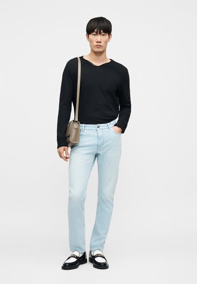 Νέος άνδρας που φοράει μαύρο μπλουζάκι με μακριά μανίκια, ανοιχτόχρωμο μπλε τζιν, μαύρα και άσπρα loafers και μια μπεζ τσάντα χιαστί, στέκεται με το ένα χέρι στην τσέπη.