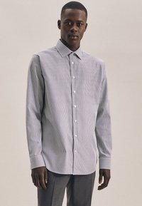 Man in een lichtgekleurde overhemd met lange mouwen en een lichte streep, met donkere broeken, staand tegen een effen neutrale achtergrond.