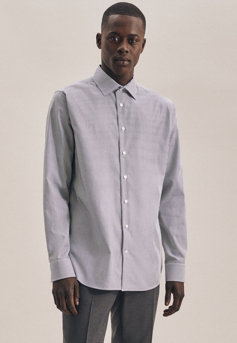 Man in een lichtgekleurde overhemd met lange mouwen en een lichte streep, met donkere broeken, staand tegen een effen neutrale achtergrond.