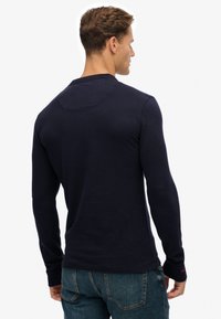 Marineblaues Langarmshirt aus einem weichen, glatten Stoff. Eng anliegendes Design mit rundem Ausschnitt und minimalen Nahtdetails. Kombiniert mit blauen Jeans aus Denim.