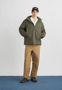 Maslinasto zelena jakna na patentni zatvarač s kapuljačom, uparen s krem polo majicom, smeđim hlačama i crnim cipelama. Jednostavan dizajn, glatka tekstura, fokus na funkcionalnost.