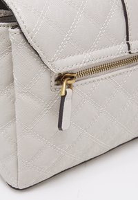 Sac à main gris clair avec un motif matelassé, fermeture éclair dorée et finition texturée. Présente un logo subtil sur le matériau.