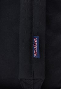 Sac à dos en tissu noir avec une étiquette de logo Jansport bleue et rouge bien visible, présentant une surface texturée et des coutures renforcées.