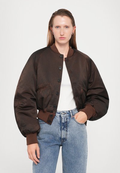 Brown bomber jacket with a satin finish, ribbed cuffs, and a button-up front. Worn over a white t-shirt and paired with blue high-waisted jeans.