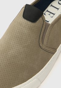 Slip-on sportcipő, perforált világosbarna felsőrésszel, fekete részlettel a sarkon és fehér gumitalppal. Simább textúrájú, minimalista dizájn.