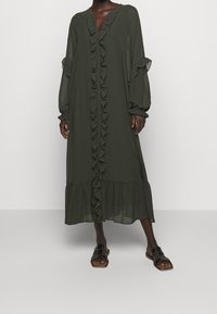 Largo vestido de gasa verde oliva con volantes en el frente, detalles de botones y mangas abullonadas. Presenta un dobladillo con volantes en capas. Combinado con sandalias planas.