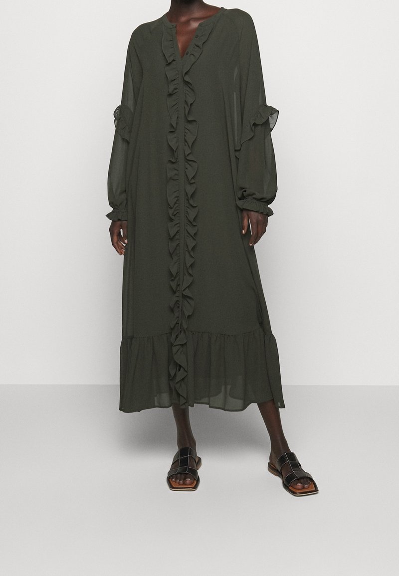 Largo vestido de gasa verde oliva con volantes en el frente, detalles de botones y mangas abullonadas. Presenta un dobladillo con volantes en capas. Combinado con sandalias planas.