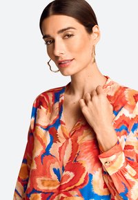 Bunte Bluse mit Blumenmuster in Rot-, Blau- und Cremetönen; weicher Stoff; V-Ausschnitt; lange Ärmel mit Knopfakzenten; kombiniert mit goldenen Creolen.