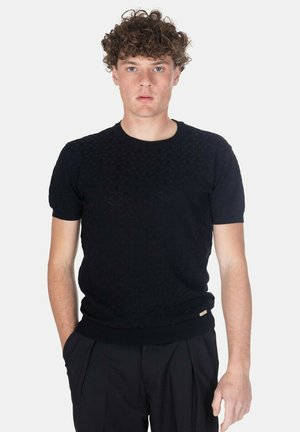Giovane uomo con capelli ricci che indossa un maglione nero a maniche corte con trama e pantaloni neri, posando con una mano in tasca.