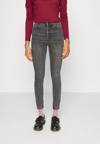 Grå jeans med hög midja, femknäppt stängning, smal passform och fransad nederkant. Bärs med svarta skor och prickiga strumpor.