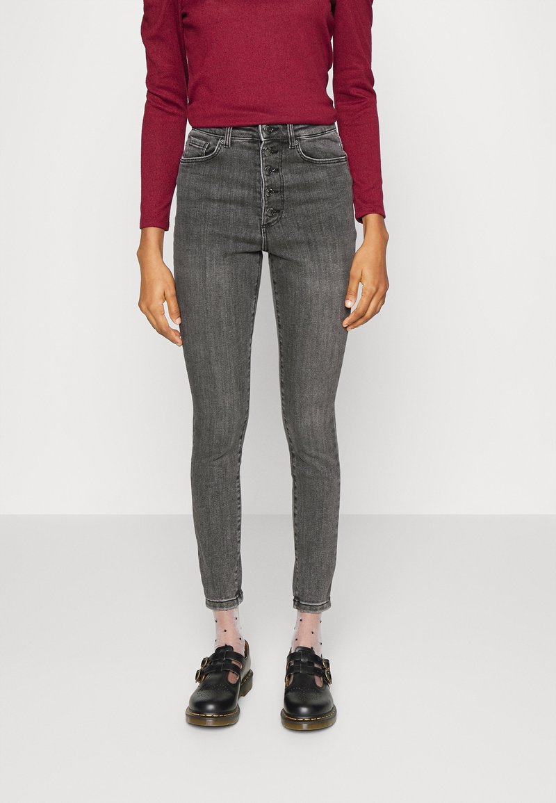 Grå jeans med hög midja, femknäppt stängning, smal passform och fransad nederkant. Bärs med svarta skor och prickiga strumpor.