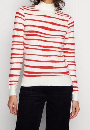 Kobieta ubrana w biały sweter z golfem z nieregularnymi czerwonymi poziomymi paskami i czarne sztruksowe spodnie, stojąca na jednolitym tle.