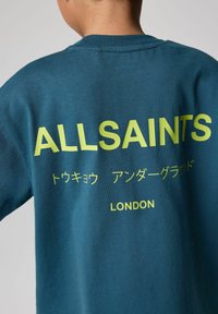 Teal sweatshirt med stort gult tekst: "ALLSAINTS," med japanske tegn nedenunder, og "LONDON" nederst. Blød tekstur.