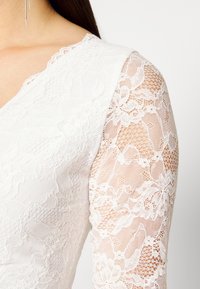 Vit spetsklänning med blommönster; långa ärmar har ett genomskinligt spetsöverdrag. Slät tyg med scalloped halsringning.