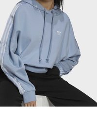 Person klädd i en ljusblå Adidas-hoodie med vita ränder på ärmarna och svarta byxor, sittande med en hand i fickan på en vit block.