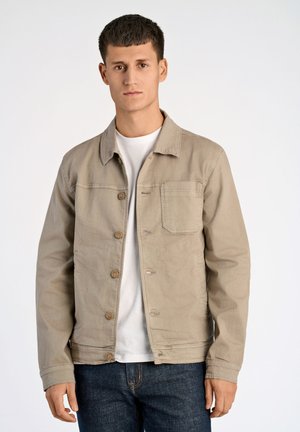 Chaqueta de mezclilla beige con cuello tipo solapa, cierre de botones y bolsillo en el pecho. Lleva puesta una camisa blanca y combinada con jeans oscuros. Textura suave.