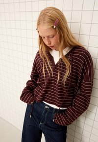 Pull-over rayé bordeaux et blanc avec capuche, doté d'une encolure boutonnée et de cordons de serrage. Un jean bleu foncé complète la tenue.