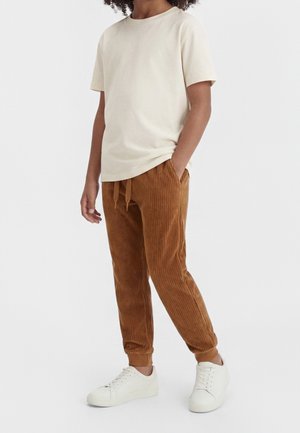 Ung person iført en ensfarvet beige t-shirt med korte ærmer, brune joggebukser i ribstof med snore og hvide sneakers, stående med den ene hånd i lommen.