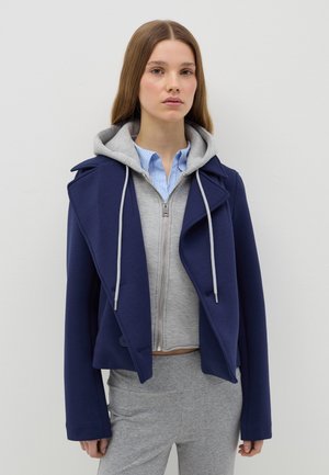 Giacca cropped blu navy con un design doppiopetto, caratterizzata da una felpa con cappuccio grigia sotto e una camicia a righe azzurre chiare.