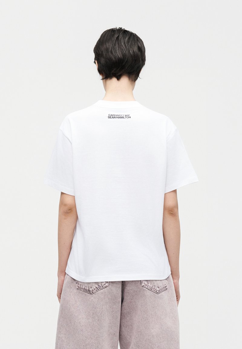 Persona con capelli corti e scuri che indossa una maglietta bianca con la scritta "Carhartt WIP Sean Hamilton" sulla parte superiore della schiena e pantaloni grigio chiaro.