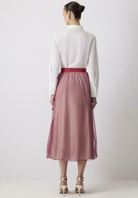 Femme aux cheveux noirs en chignon portant un chemisier blanc à manches longues, une jupe midi en voile de rose poussiéreux, et des sandales à talons blancs, de dos.