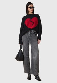 Schwarzer oversized Pullover mit einem strukturierten roten Herzdesign, kombiniert mit grauen High-Waist-weit geschnittenen Jeans und schwarzen spitzen Stiefeletten.