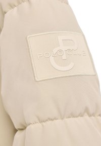 Beżowa pikowana tkanina z przeszytym prostokątnym łatą, na której znajduje się wytłoczone nakładające się litery "P" i "C" oraz tekst "POLO CLUB".