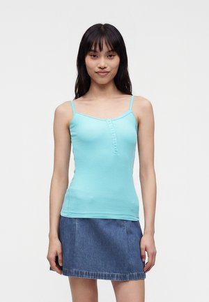 Jeune femme aux longs cheveux foncés, portant un haut sans manches bleu clair boutonné et une jupe en jean bleue, debout devant un fond uni.