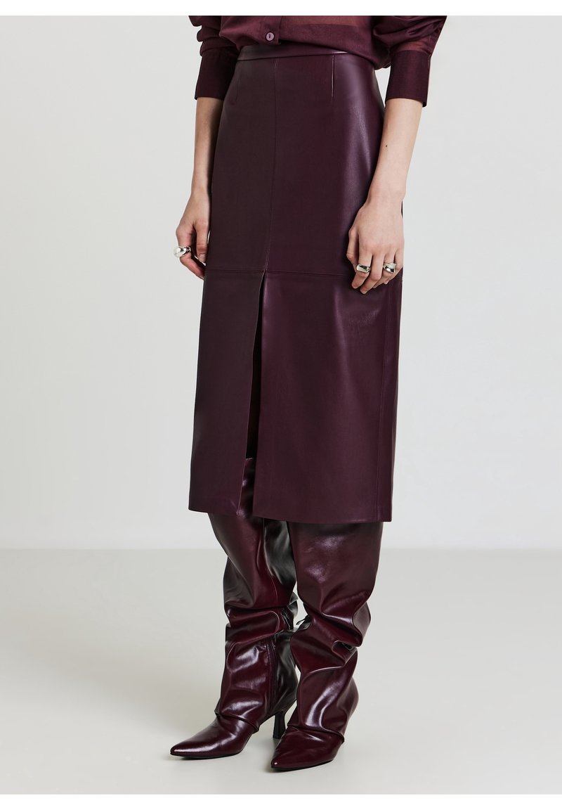 Femme portant une jupe midi en cuir bordeaux avec une fente avant et des bottes à talons larges assorties, debout devant un fond uni.