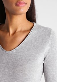 Maglione grigio con scollo a V realizzato in tessuto lavorato a maglia leggero, caratterizzato da un design aderente e una texture liscia, con dettagli di cuciture visibili attorno al collo.