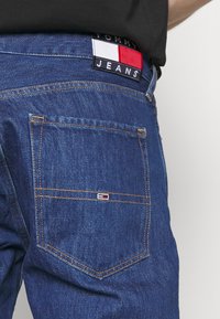 Modré džíny z denimu s viditelným zadním kapsou, na kterém je značková nášivka v červené, bílé a modré barvě s prošitými detaily.