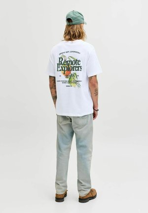 Muž s dlhými vlasmi, ktorý má na sebe svetlozelenú čiapku, biele tričko s nápisom "Remote Explorers" a grafikou ovocia, svetlé rifle a hnedé topánky, stojaci chrbtom.