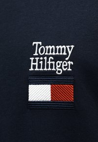 Υφασματό βαμβακερό σε ναυτικό μπλε με κεντημένο το "Tommy Hilfiger" σε λευκό, συνοδευόμενο από ριγέ λογότυπο σε κόκκινο, λευκό και μπλε με λεπτομέρειες υφής.