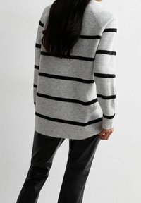 Kobieta z długimi czarnymi włosami ubrana w szary sweter w czarne poziome paski oraz czarne spodnie, przedstawiona z tyłu.